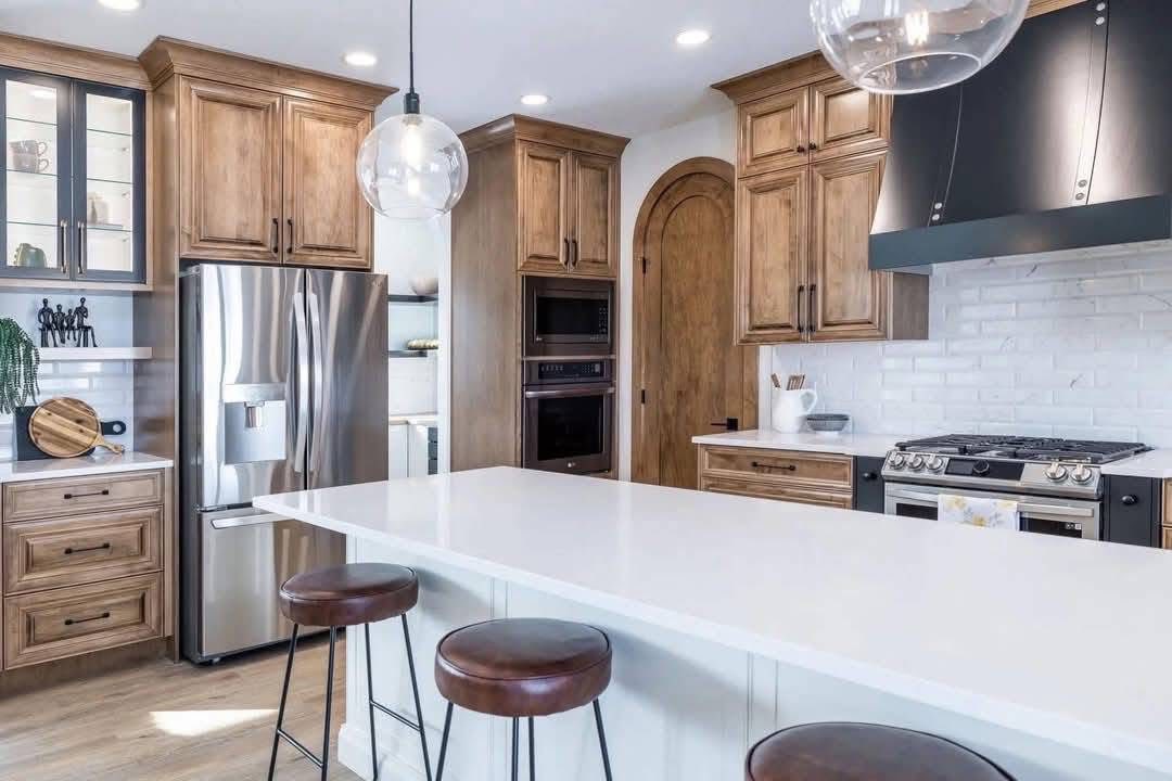 Modern kitchen with white countertops and wooden cabinets. Features stainless steel appliances, bar stools, pendant lights, and a cozy, inviting tone.