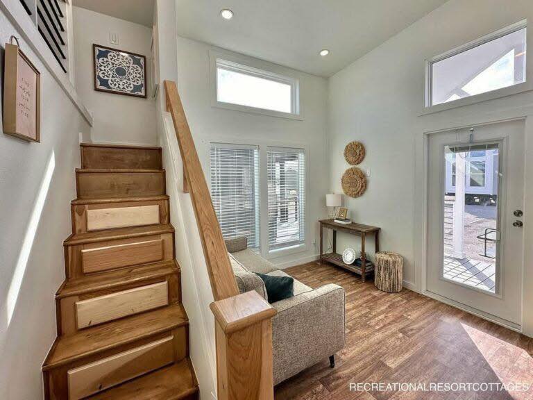 Cozy living area with wooden stairs, light wooden floors, and a beige sofa. Sunlight streams through tall windows, creating a warm and inviting atmosphere.