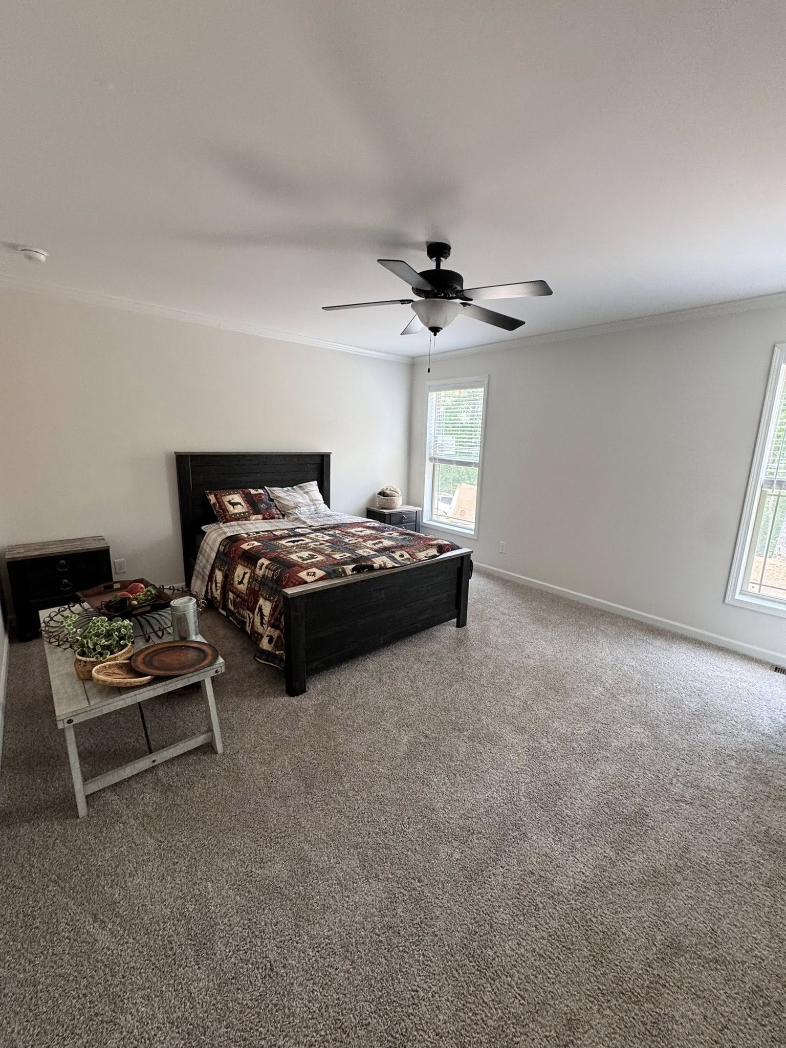 A cozy bedroom with a large dark wooden bed, patterned quilt, and a bedside table. Carpeted floor, ceiling fan, and natural light from a window.