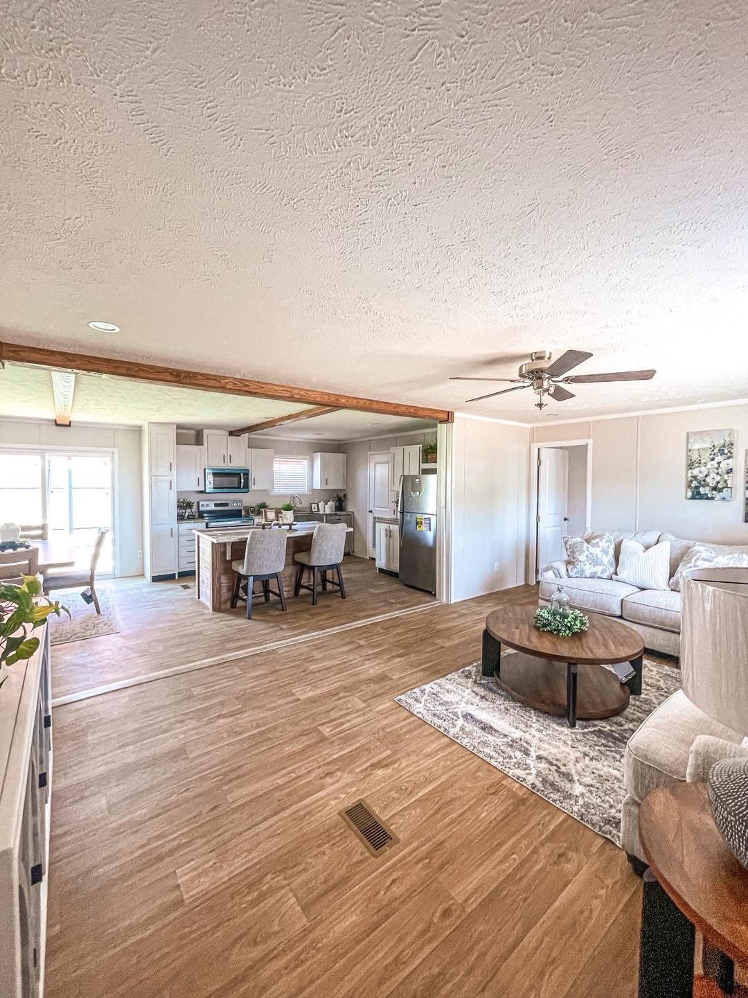 Spacious open-plan living room and kitchen with wooden floor, ceiling fan, cozy white sofa, round coffee table with plant, and modern kitchen island.