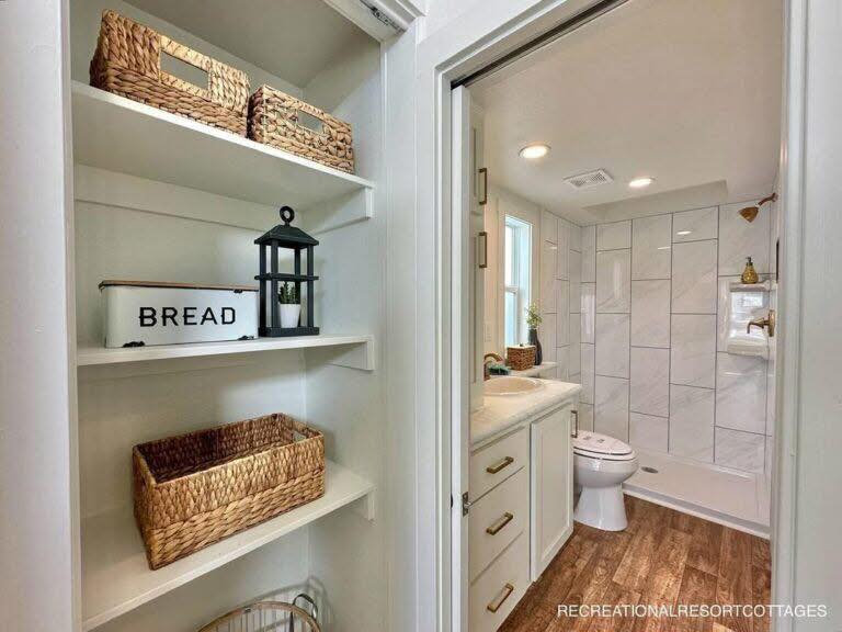 Narrow bathroom with white cabinets, modern tiles, and wooden flooring. Shelves with woven baskets and a white "BREAD" box are visible in the foreground.
