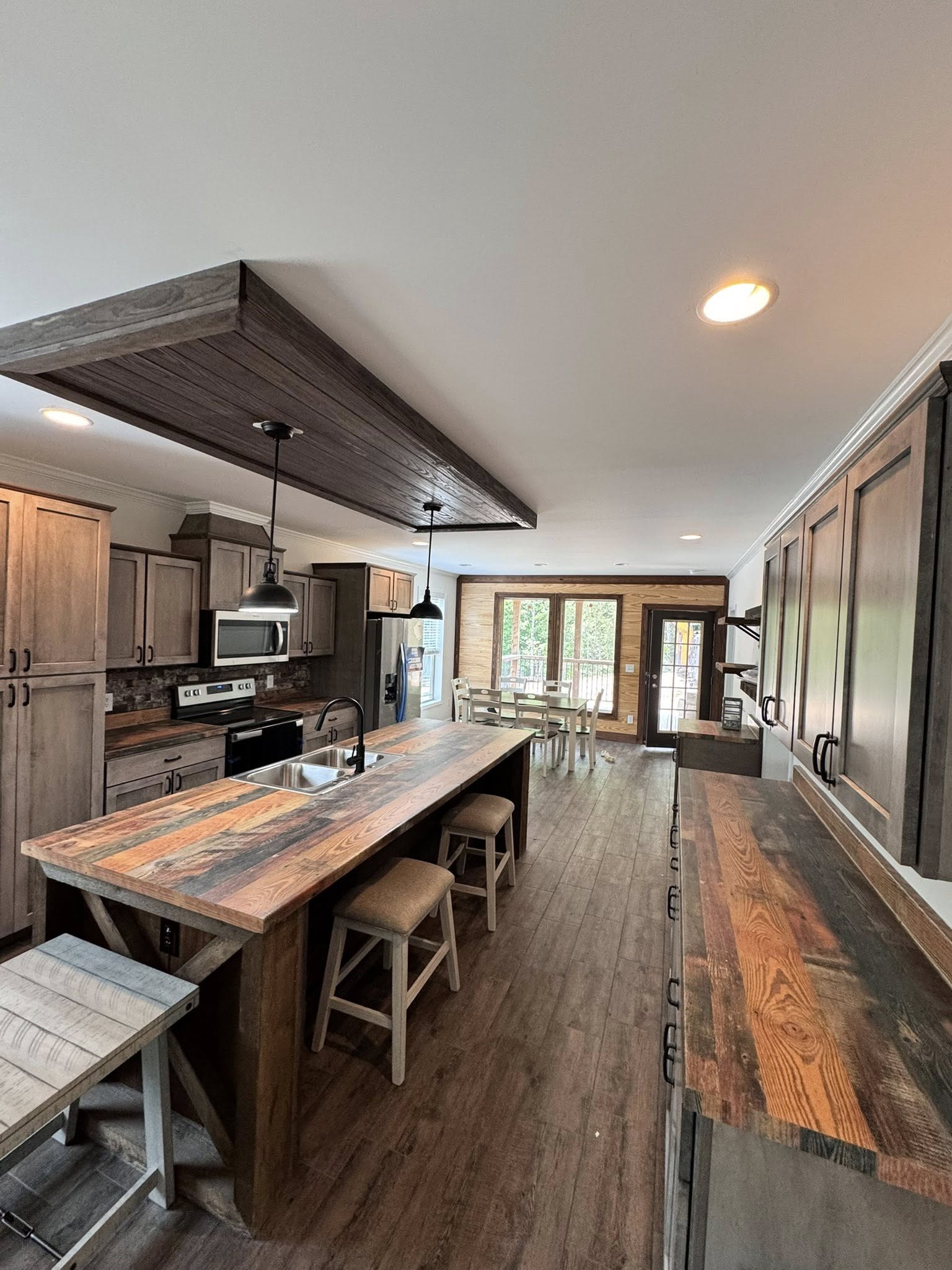 Spacious rustic kitchen with wooden countertops and cabinets, center island with stools, hanging pendant lights, and large windows in the background.