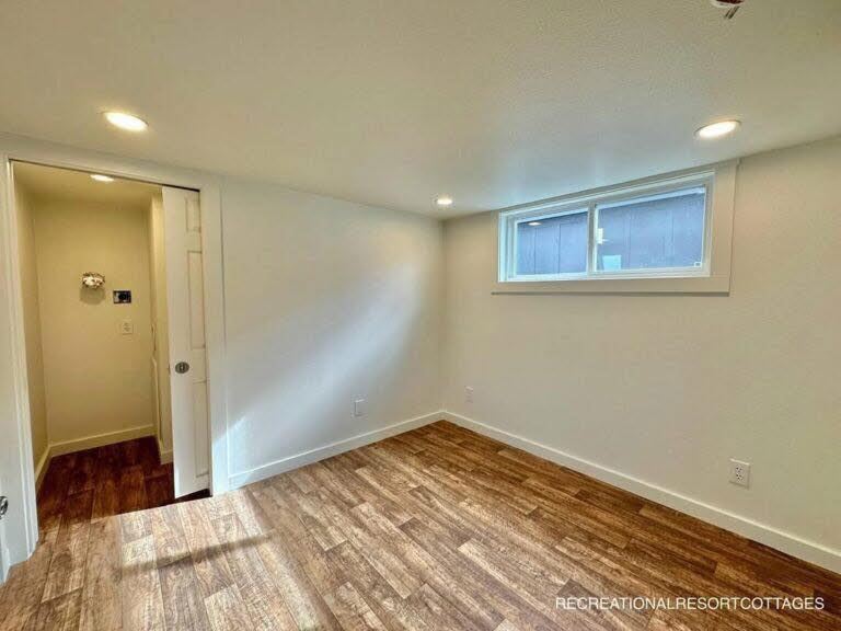 A small, bright room with light wood flooring, white walls, and ceiling with recessed lighting. A narrow window provides natural light. A door leads to a hallway.