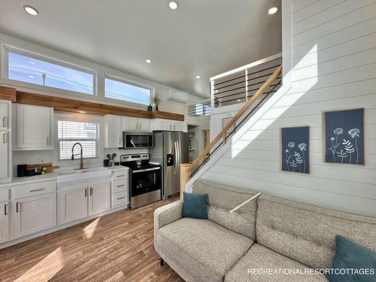 Modern tiny house interior with open layout. Bright kitchen features white cabinets, stainless steel appliances, and wood accents. Cozy living area includes a gray sofa and blue pillows. Sunlight streams through high windows, illuminating the space and creating a warm, inviting atmosphere.