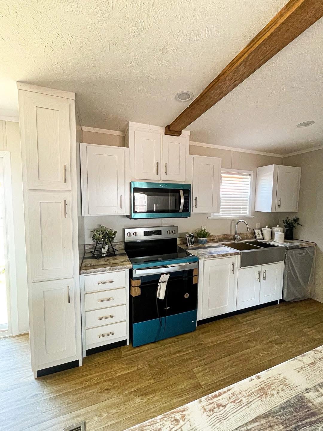 Modern kitchen with white cabinets, a wooden beam ceiling, and wood flooring. Stainless steel oven and microwave beneath a large window. Bright and clean.