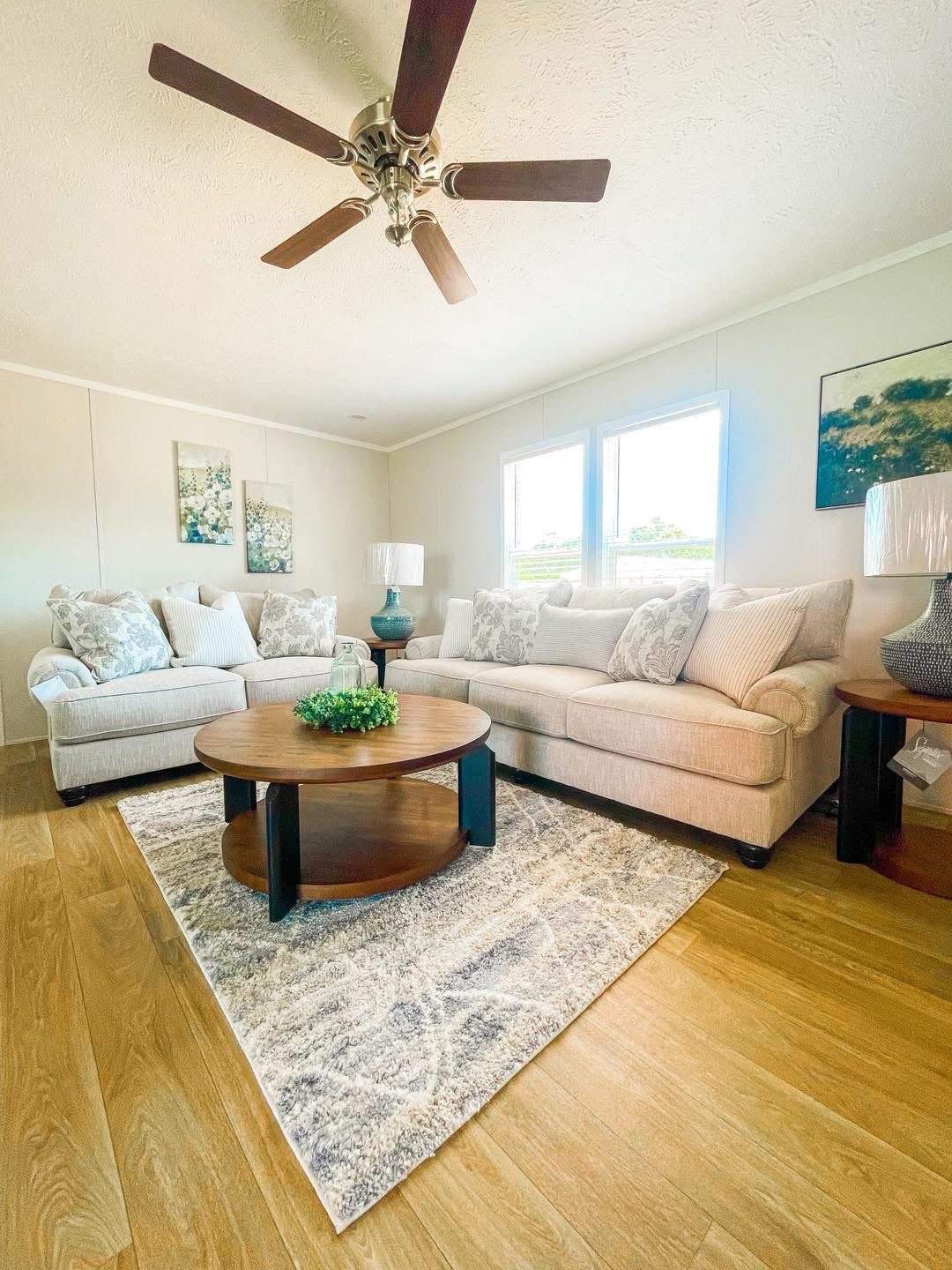 Spacious living room with beige sofas, patterned cushions, and a round wooden coffee table on a textured rug. Large windows and ceiling fan add brightness.