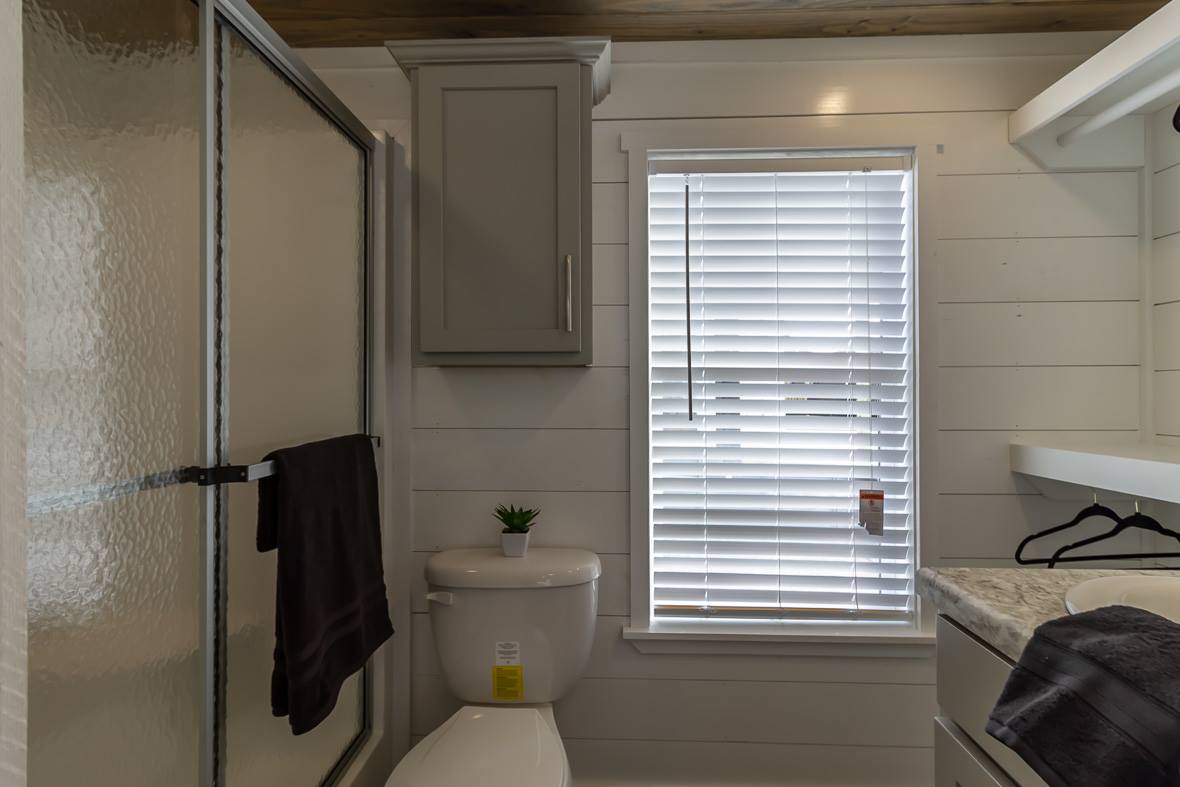 Compact bathroom with a glass shower door, white shiplap walls, and a window with blinds. Gray cabinet above a toilet with a small plant.