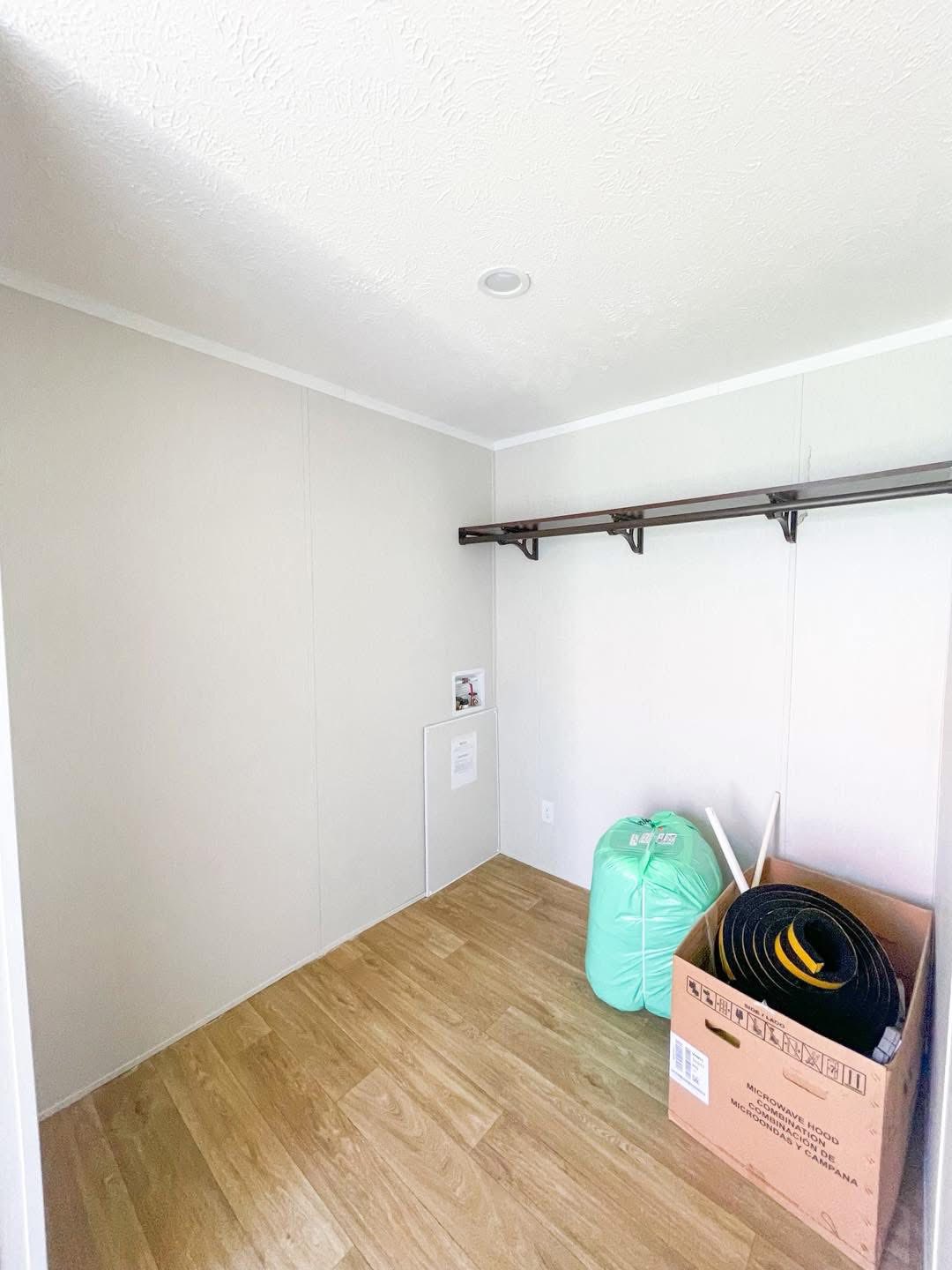 A small, empty closet with light beige walls and wood flooring. A wooden shelf runs along one wall. A cardboard box and green bag are on the floor.