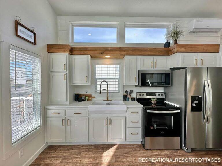 Modern kitchen with white cabinets, a farmhouse sink, stainless steel appliances, and wood accents. Large windows allow abundant natural light.