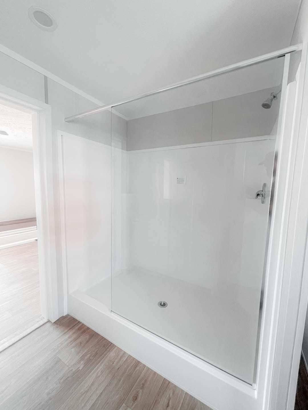 A modern, minimalist shower with clear glass doors, white walls, and light wooden flooring. The space conveys a clean, serene atmosphere.