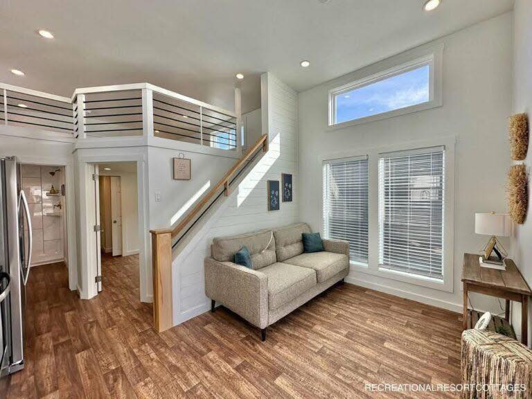 Bright and cozy living room with vaulted ceiling and large windows. Beige sofa, wooden floors, staircase to loft, modern decor, and ample natural light.