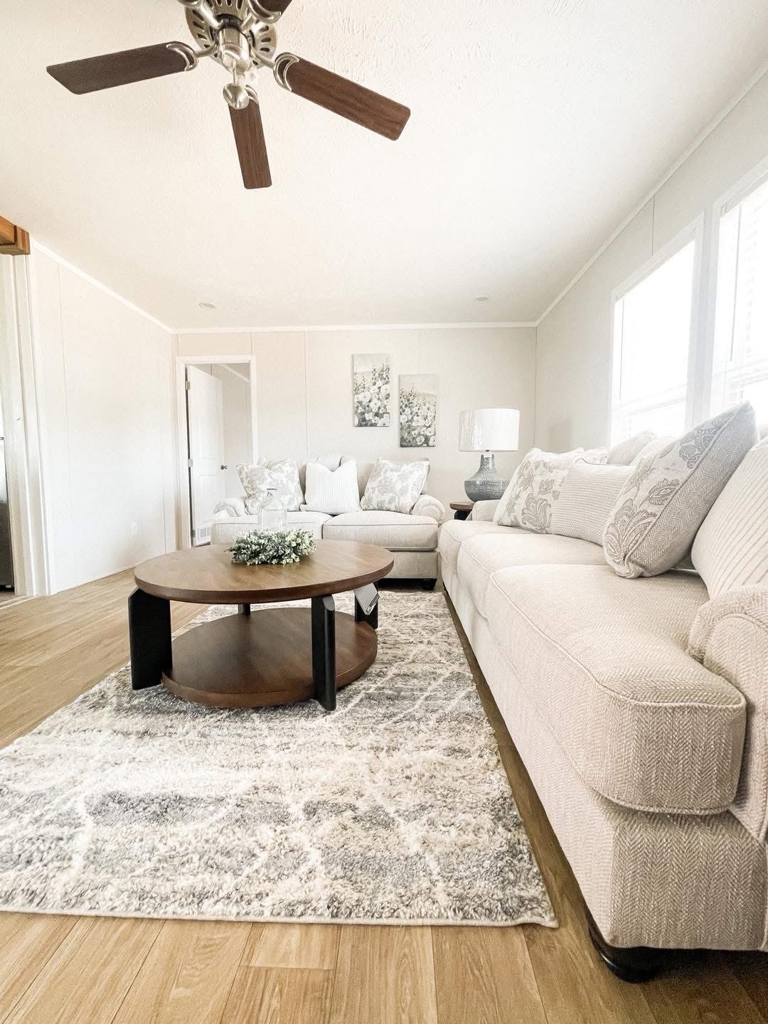 Bright living room with beige sofas and floral cushions. A wooden coffee table with greenery sits on a gray patterned rug. Ceiling fan above. Airy, serene feel.