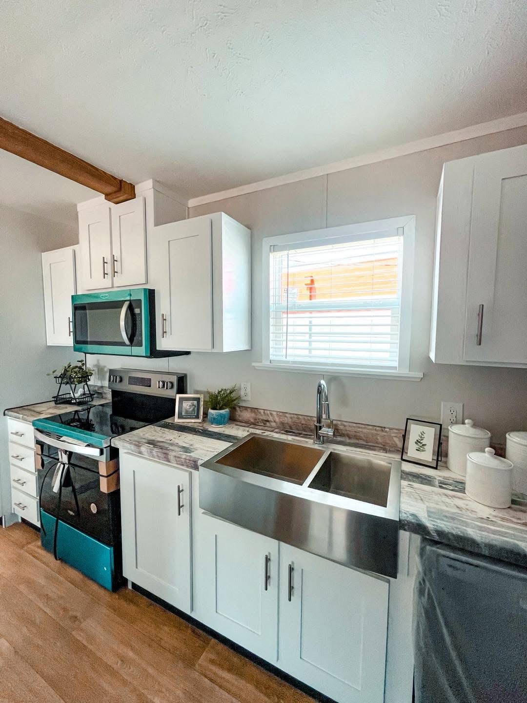 Modern kitchen with white cabinets and stainless steel appliances, including a teal oven and microwave. Marble countertop with a double sink, hardwood floor, and a window above the sink.