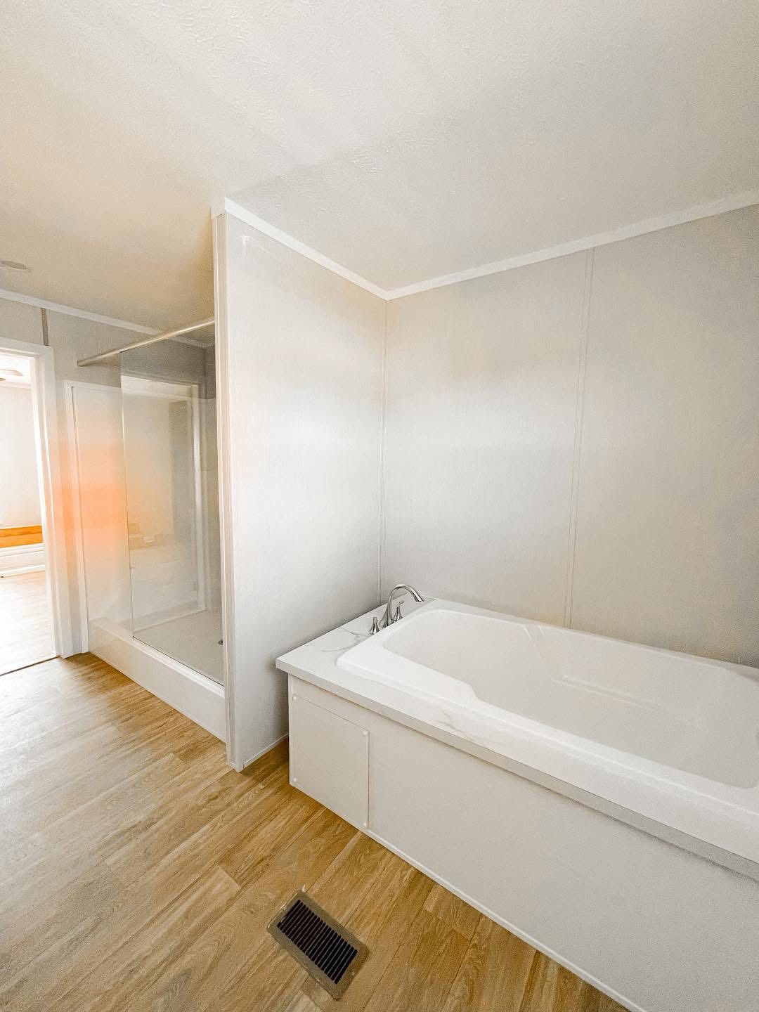 Minimalist bathroom with a white bathtub and shower, wood-style flooring, and a warm, clean ambiance. Light streams in from a partially open door.