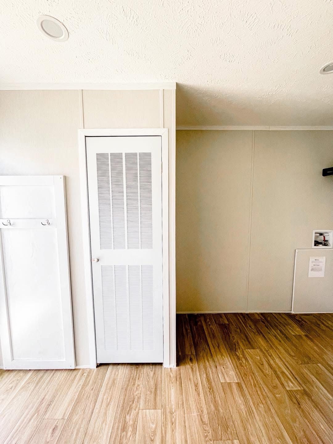 Simple interior with white louvered door and hooks on left wall. Light wood flooring, beige walls, and a cozy, minimalistic atmosphere.