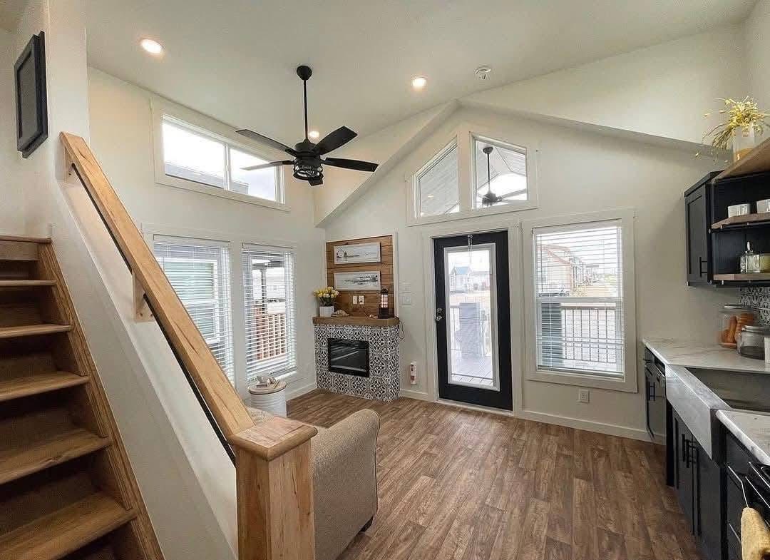 Modern tiny house interior with a high ceiling, ceiling fan, large windows, and a loft staircase. Cozy vibe with wooden floors and a small fireplace.