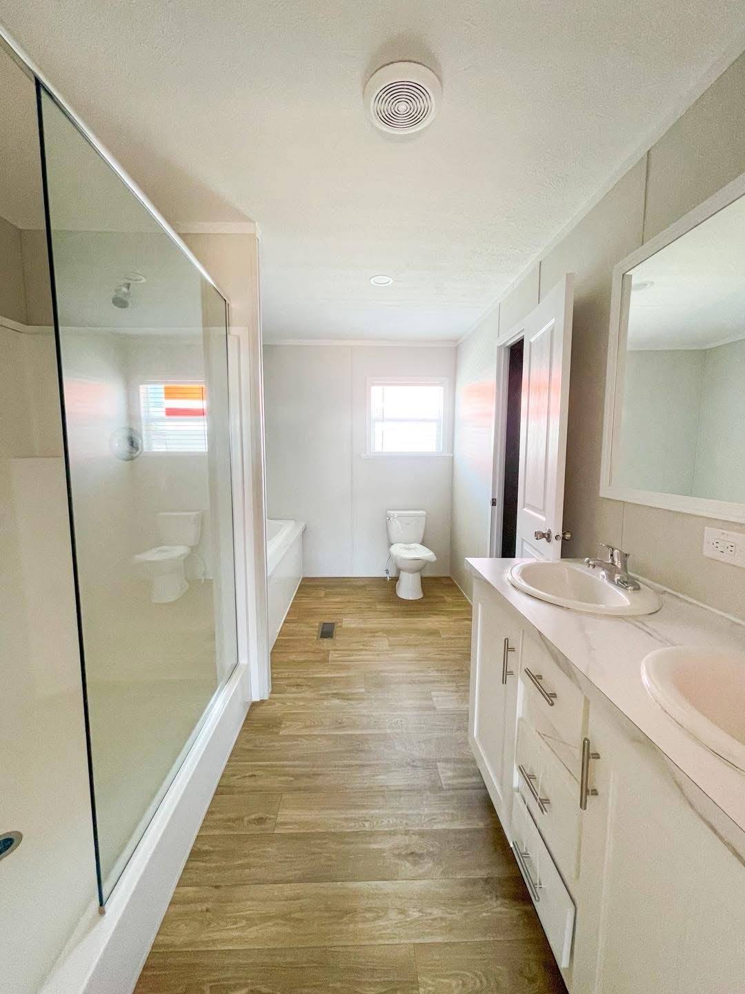 Modern bathroom with wooden floors, a glass shower on the left, a white toilet beneath a window at the back, and a double sink vanity on the right. Neutral tones create a clean, airy feel.
