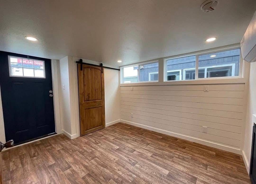 A small room with wood-patterned flooring, white shiplap walls, and a black door. A wooden sliding barn door is on the left. Natural light from narrow windows.