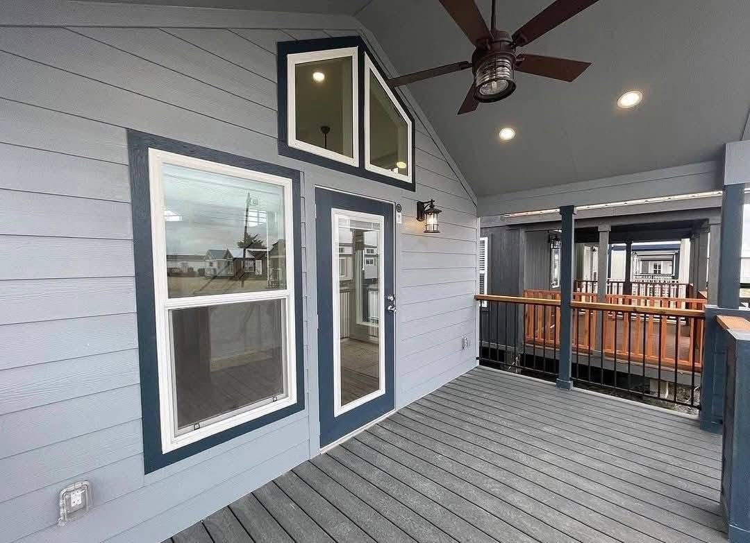 Covered porch with gray siding and a dark blue door leading inside. Features a ceiling fan, natural wood railing, and nearby homes visible outside.