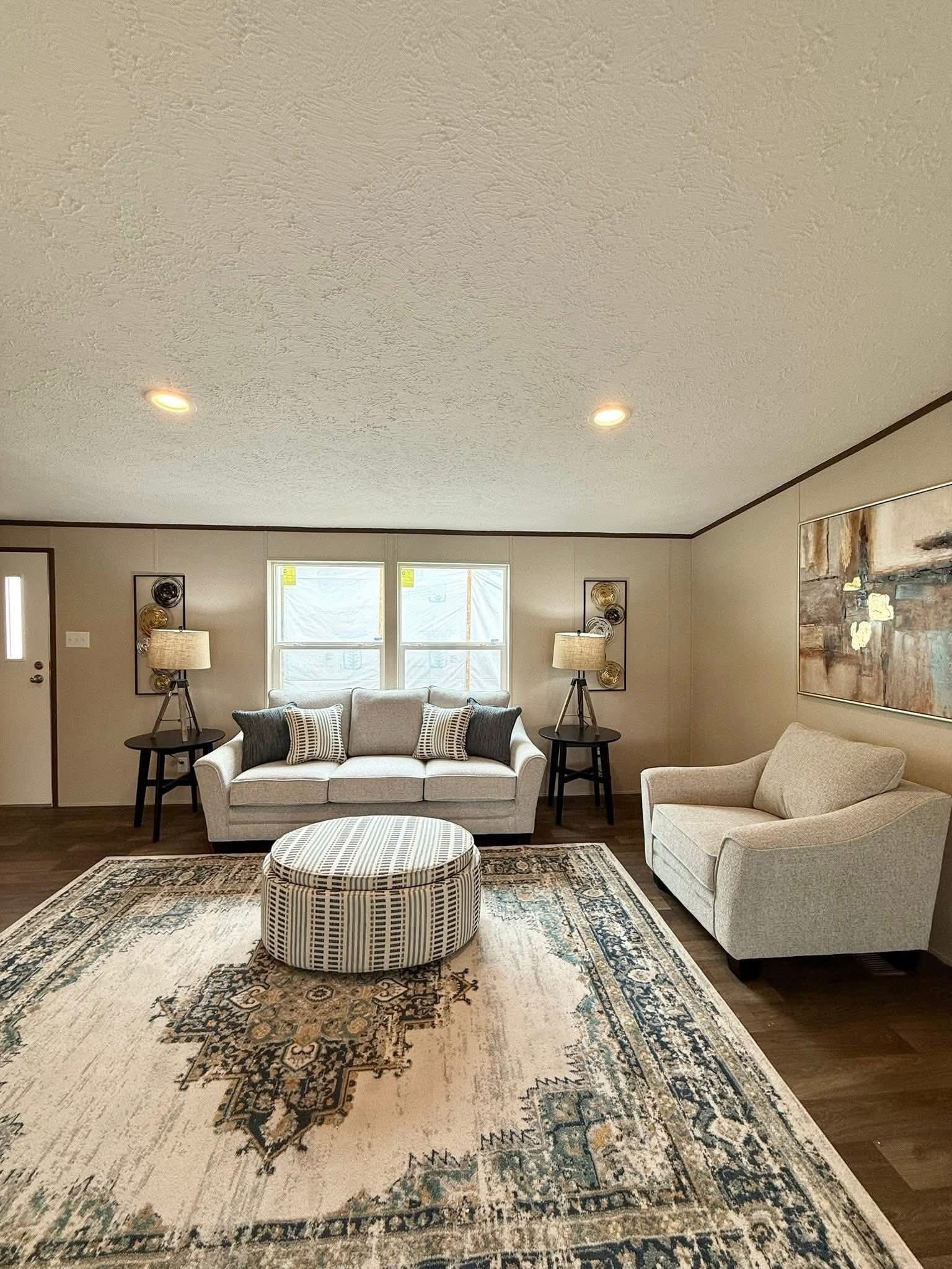 Cozy living room with a beige sofa and armchair, striped ottoman, patterned rug. Two lamps beside the sofa and abstract art on the wall, creating a warm ambiance.