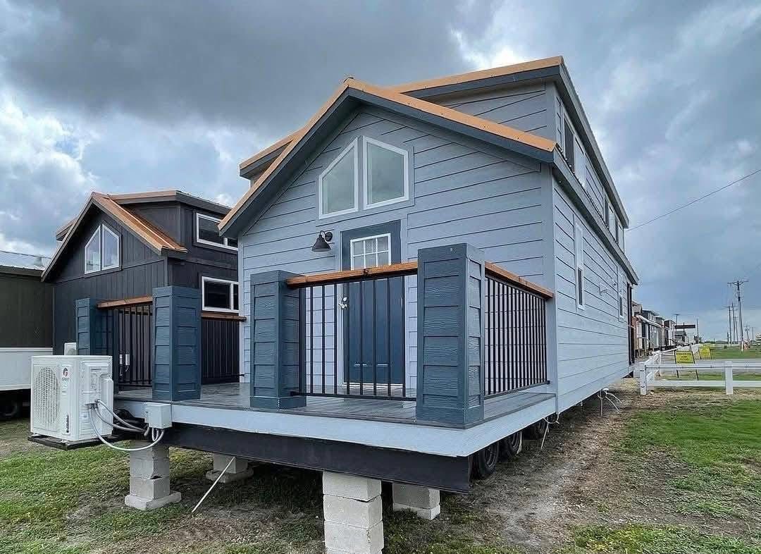 A modern tiny house on a raised platform with blue-gray siding and a small front porch. The sky is overcast, creating a moody atmosphere.