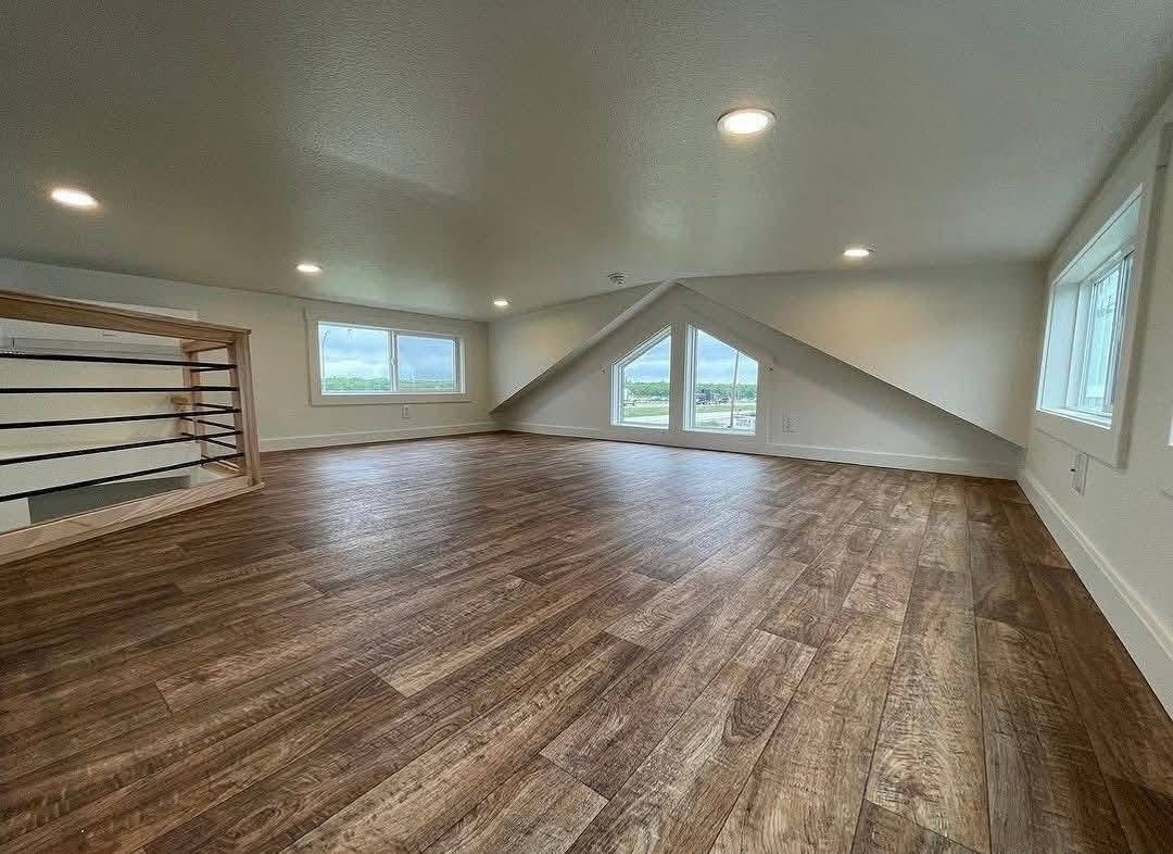 Spacious attic with wood flooring, sloped ceiling, large triangle window overlooking a scenic view, and recessed lighting, creating an airy ambiance.