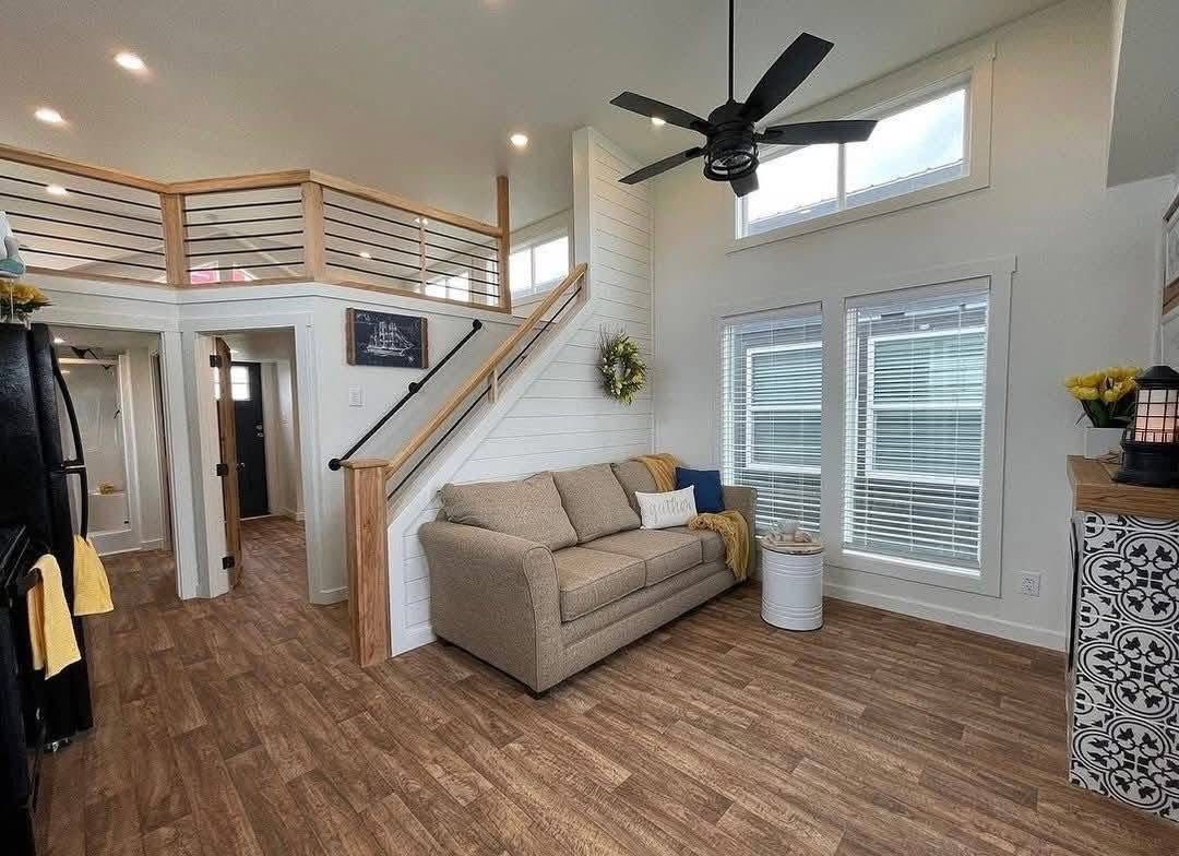 Cozy living room with a beige couch and decorative pillows, wooden floors, and a white staircase. Large windows allow natural light, creating an inviting atmosphere.