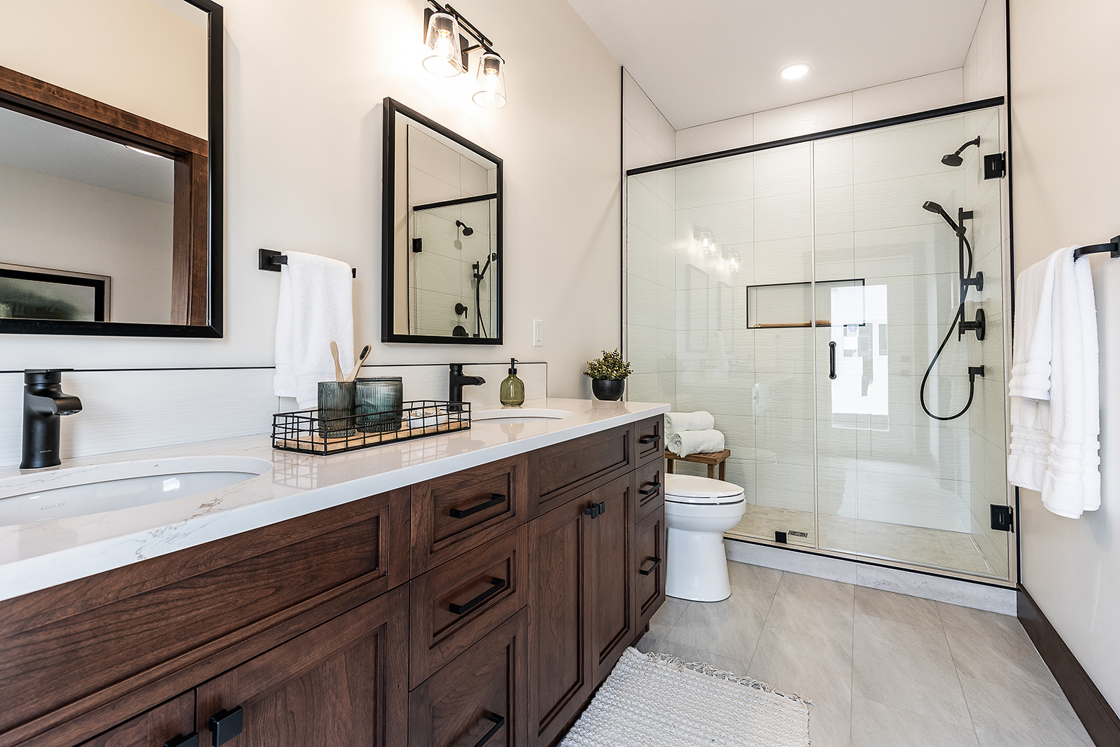 Modern bathroom with a large glass shower, dark wood vanity, double sinks, black fixtures, and rectangular mirrors. Soft lighting adds a calm ambiance.