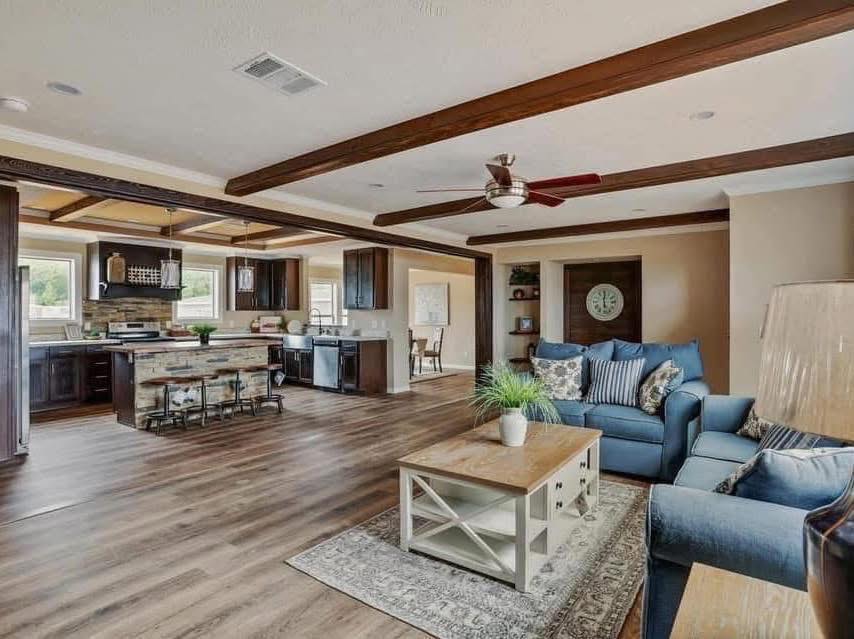 Spacious open-plan living area with wooden floors and ceiling beams. Cozy blue sofas, striped cushions, wooden coffee table with plant, and modern kitchen in background.