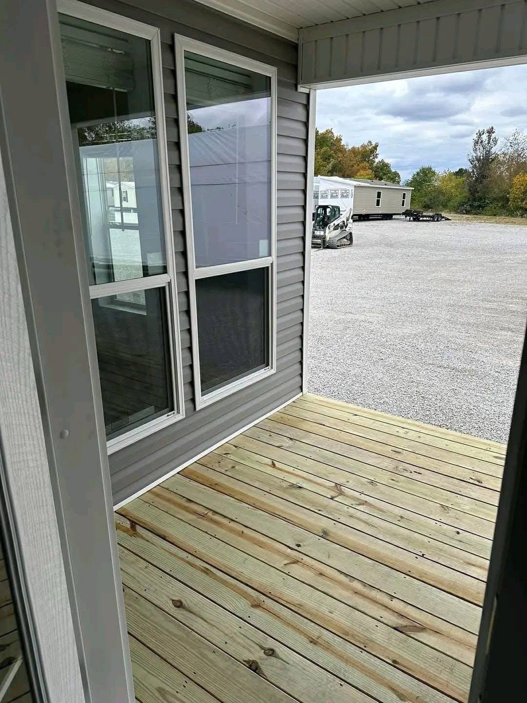 A small, covered wooden porch with smooth planks leads to a gravel lot under a cloudy sky. A modern building is visible beyond the porch.