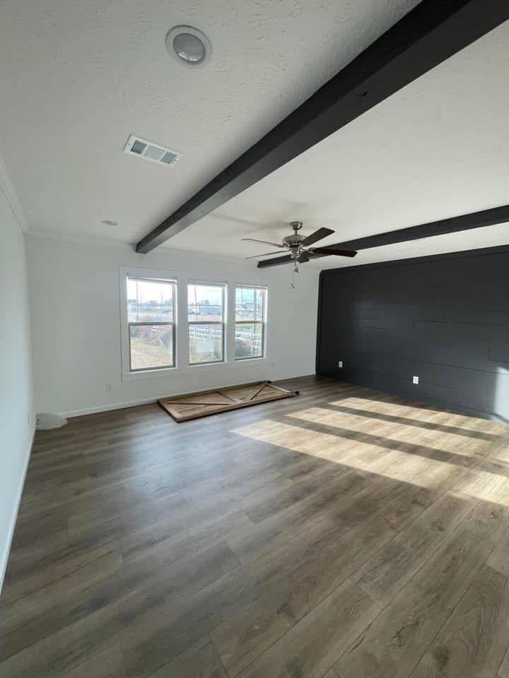 Spacious room with light wood flooring, three large windows, and dark ceiling beams. A ceiling fan is overhead, and a dark accent wall adds contrast.