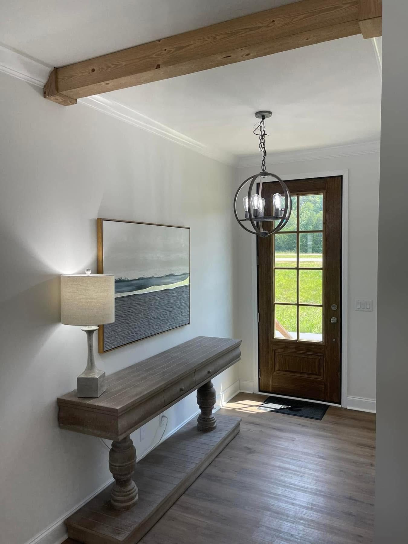 A bright entryway with a wooden console table, a table lamp, and a nature painting on the wall. A glass pendant light hangs above, near the door.