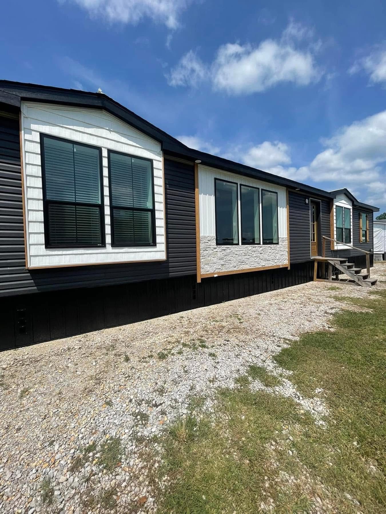 A modern, single-story mobile home with dark siding and white trim. Large windows and a grassy lawn under a bright, blue sky convey a welcoming vibe.