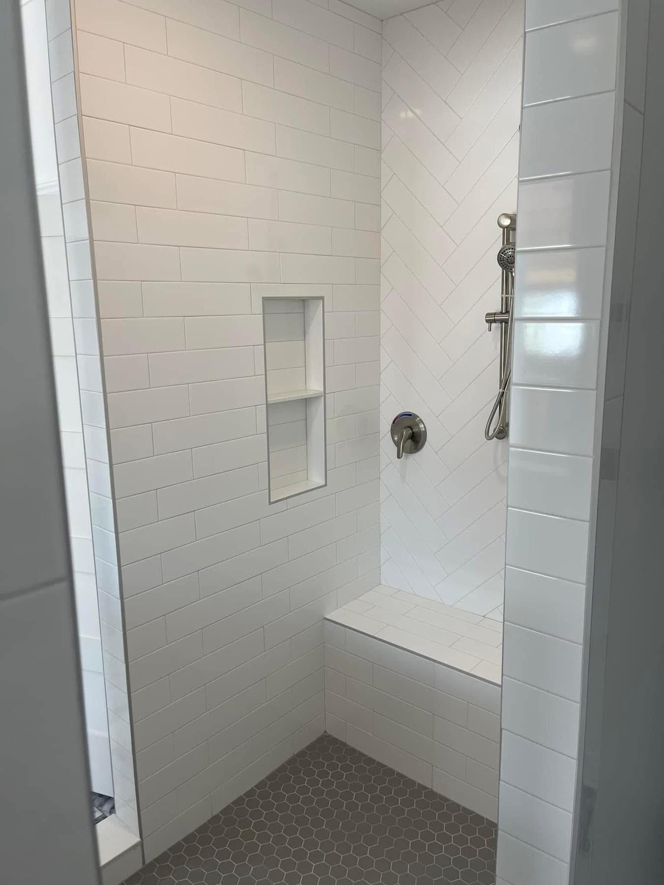 A modern shower features white subway tile walls with a herringbone pattern, a built-in shelf, and a bench. The floor has small gray hexagonal tiles.