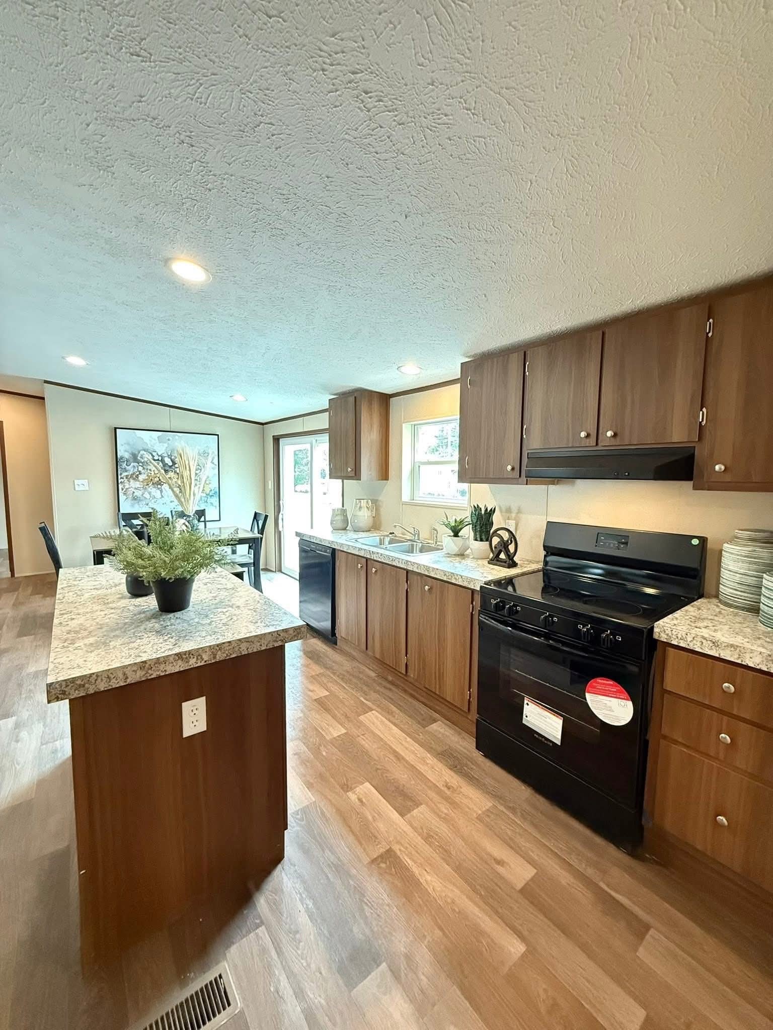 Modern kitchen with wooden cabinets, granite countertops, and a black stove. A dining area with art is visible in the background. Bright, airy ambiance.