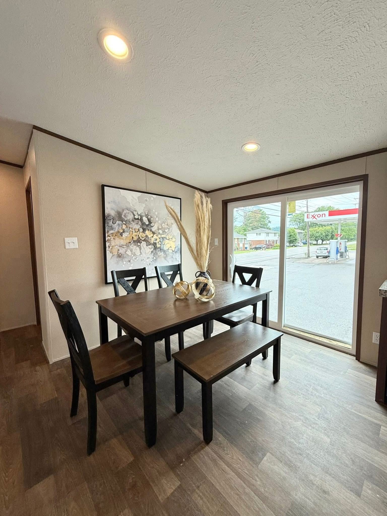 Dining area with a wooden table, chairs, and a bench. Abstract art on the wall and decorative vases on the table. Large window shows a street view.