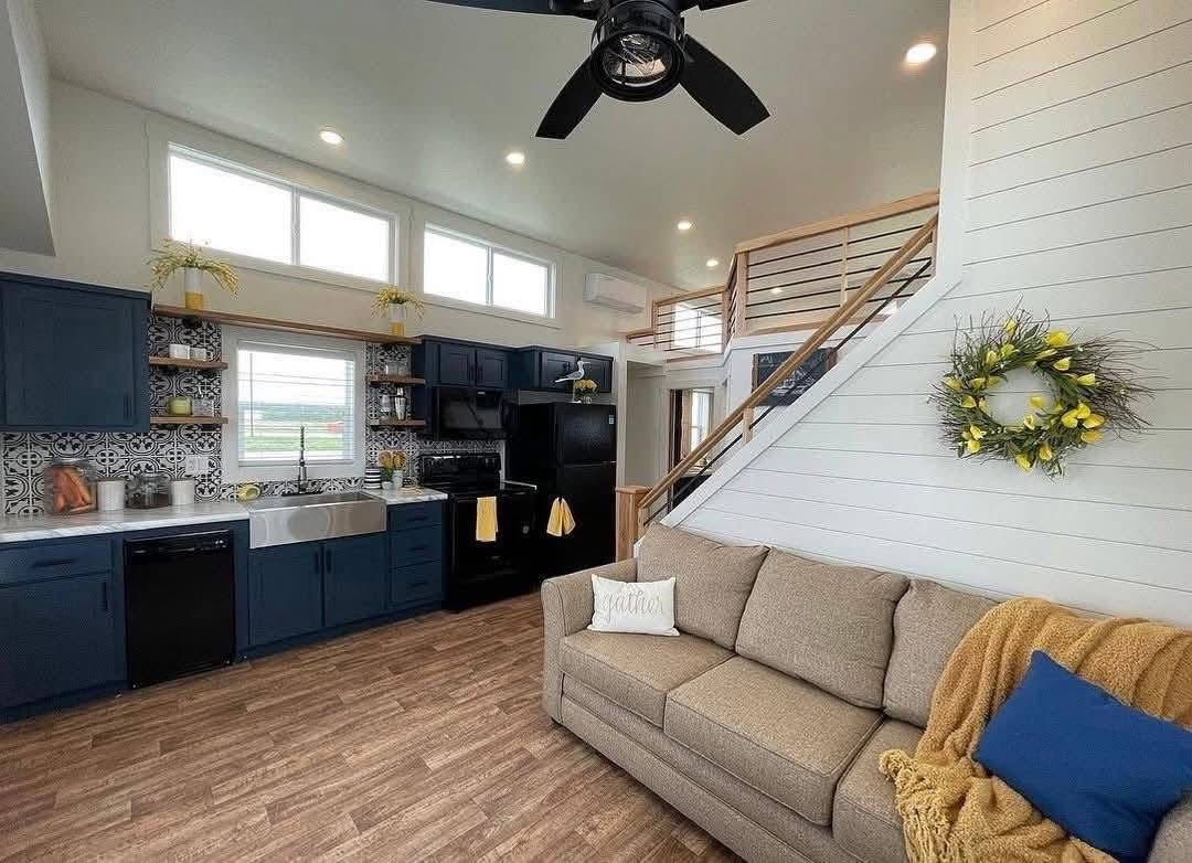 Modern living space with navy kitchen cabinets, patterned backsplash, and stainless-steel sink. Beige couch, cozy pillows, and yellow accents create a warm, inviting atmosphere.