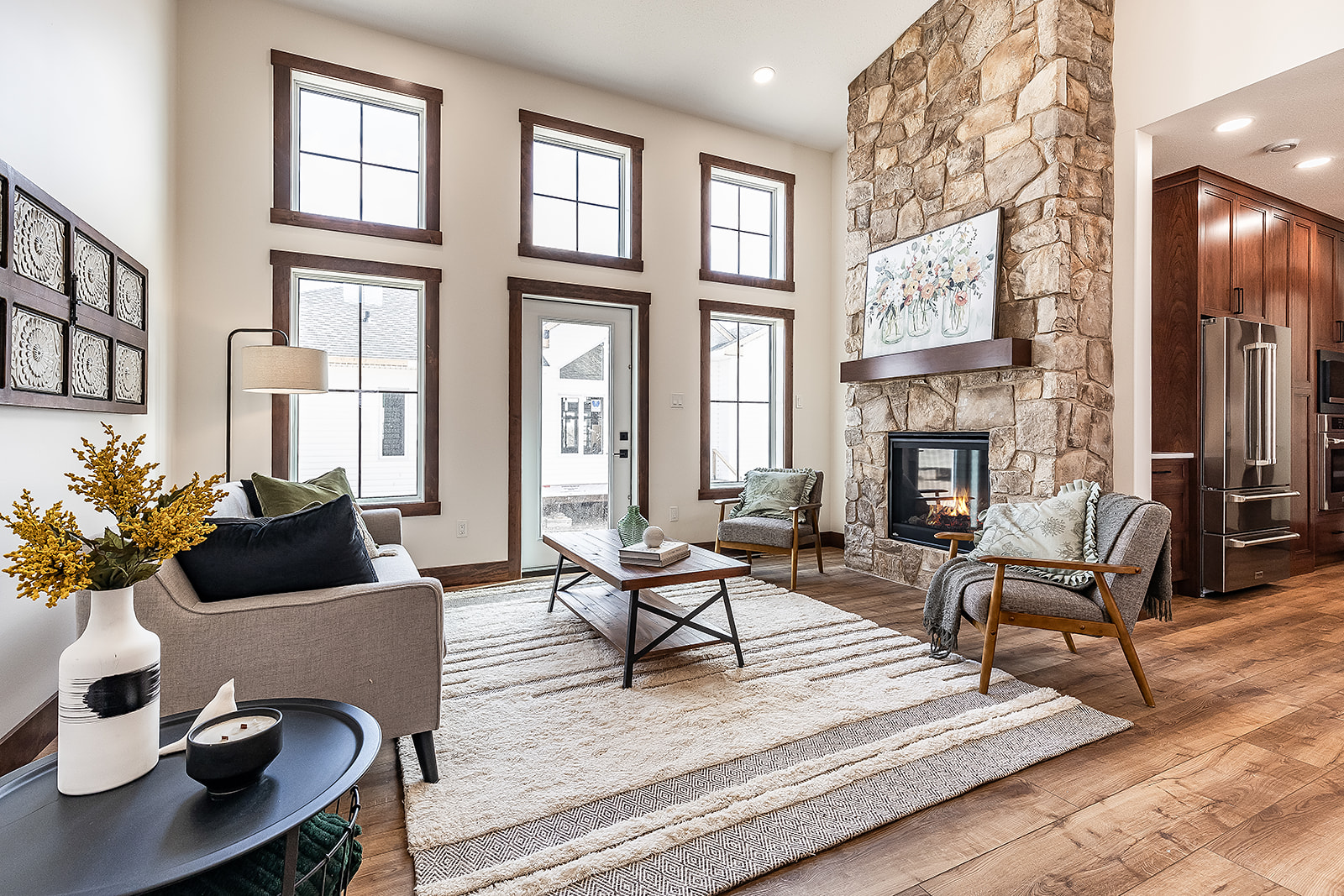 Living room with tall windows and a stone fireplace, featuring a grey sofa, wooden chairs, a neutral rug, and decorative accents. Cozy and bright ambiance.