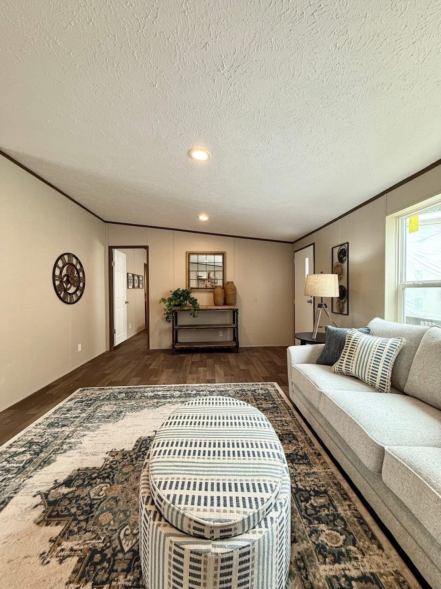 Modern living room with neutral tones, featuring a gray sofa with patterned pillows, striped ottoman, and a textured rug. A console with decor is against the wall.
