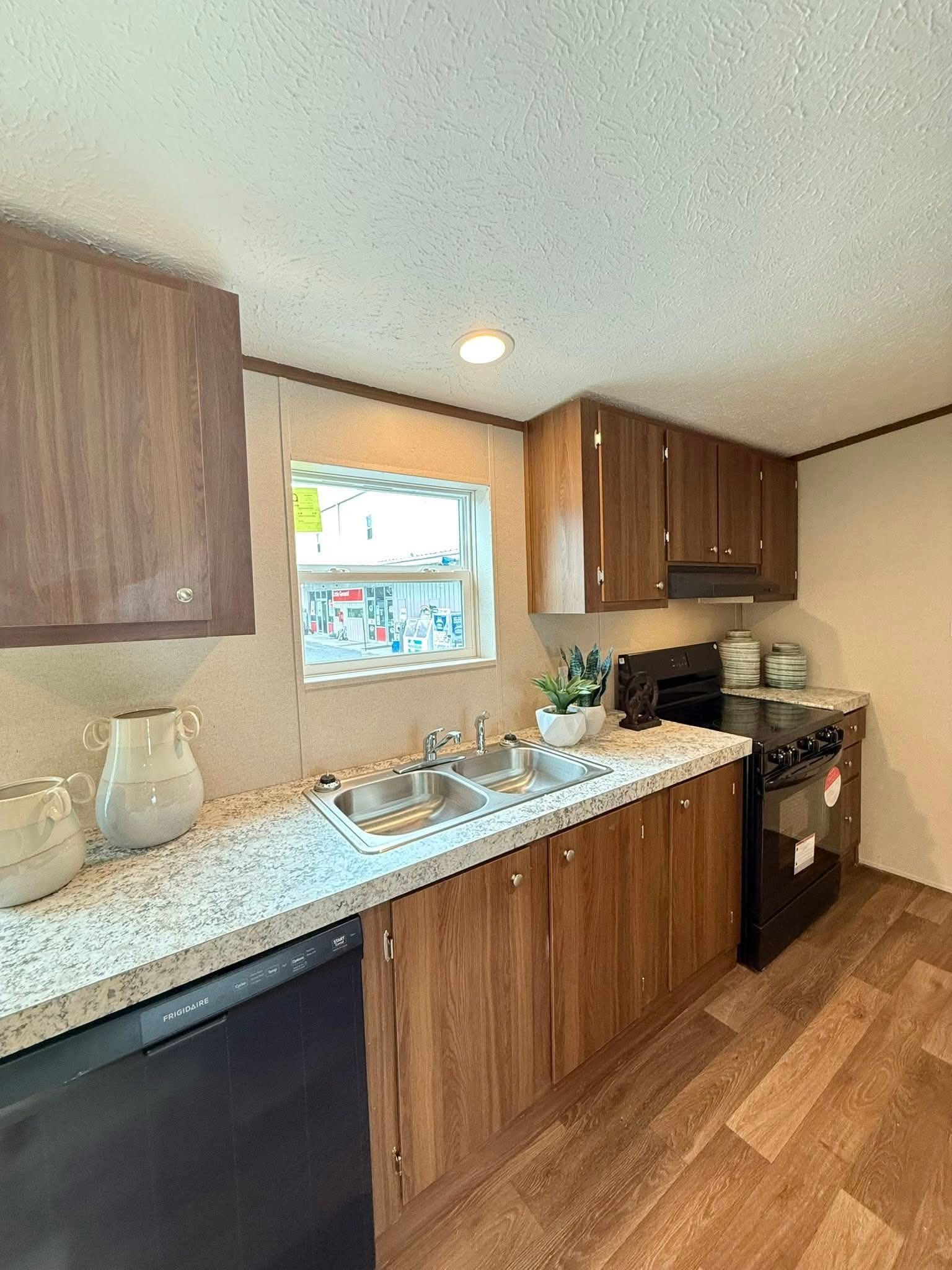 Modern kitchen with wood cabinets, granite countertop, and stainless steel double sink. Window with natural light, ceramic vases, and a potted plant add warmth.