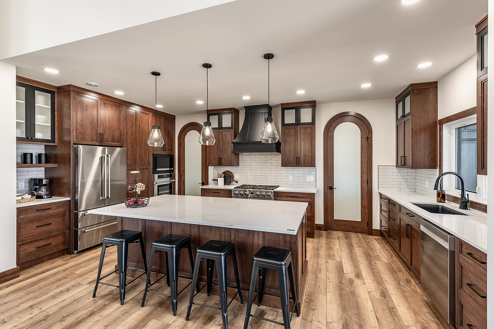 Spacious kitchen with dark wood cabinets, a large island with black stools, stainless steel appliances, pendant lighting, and a hardwood floor.