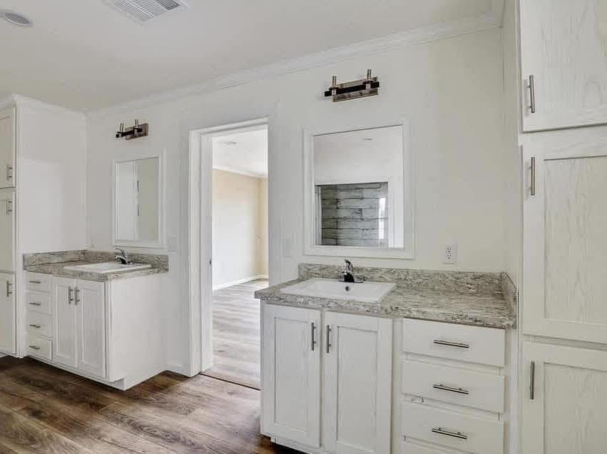 Bright, modern bathroom with dual white vanities, granite countertops, wooden flooring, large mirrors, and bright lighting. Doorway leads to adjacent room.