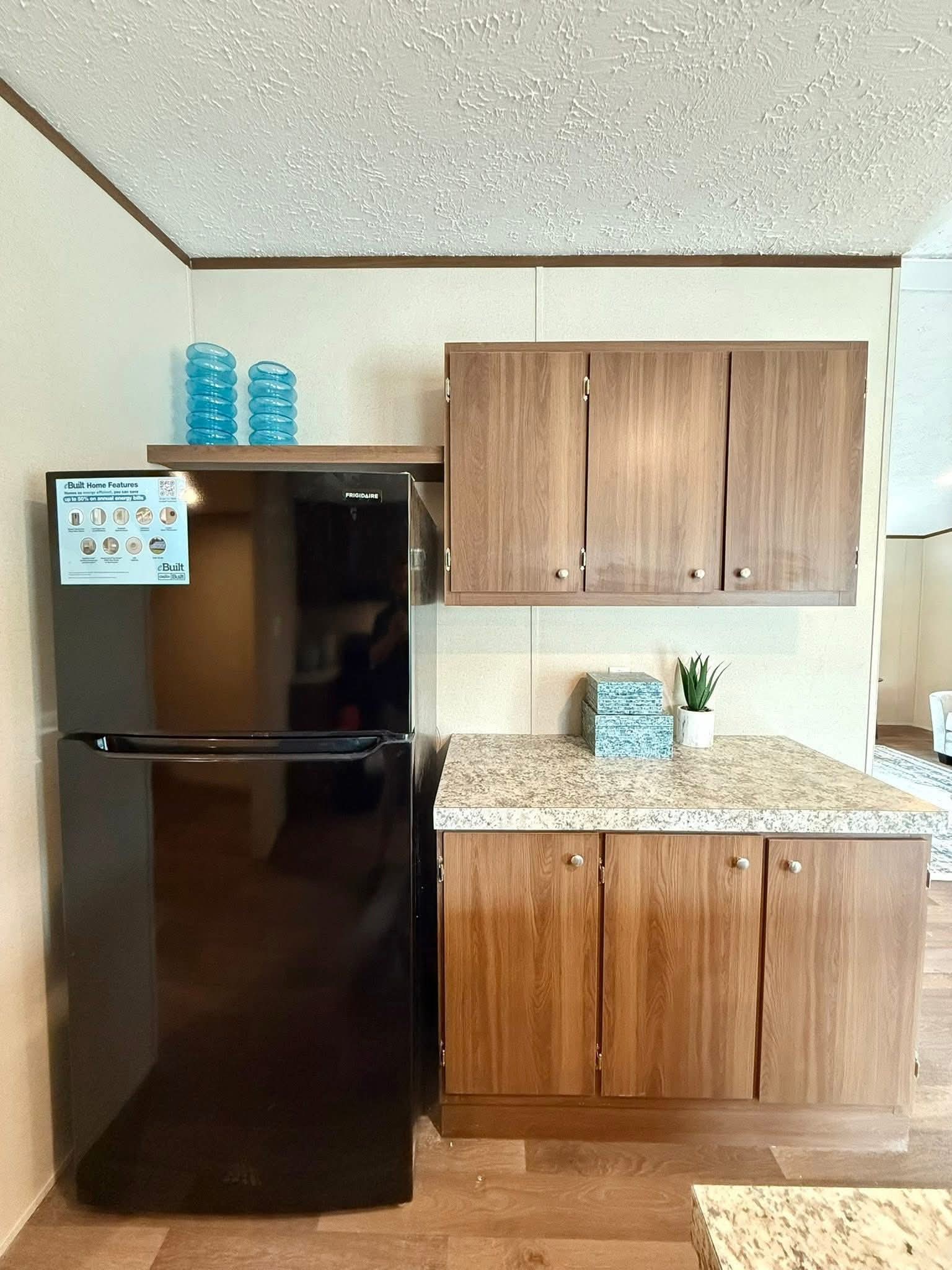 Kitchen scene with wood cabinets, a black refrigerator, and a granite countertop. Decor items include blue glass jars and a small plant, creating a cozy tone.