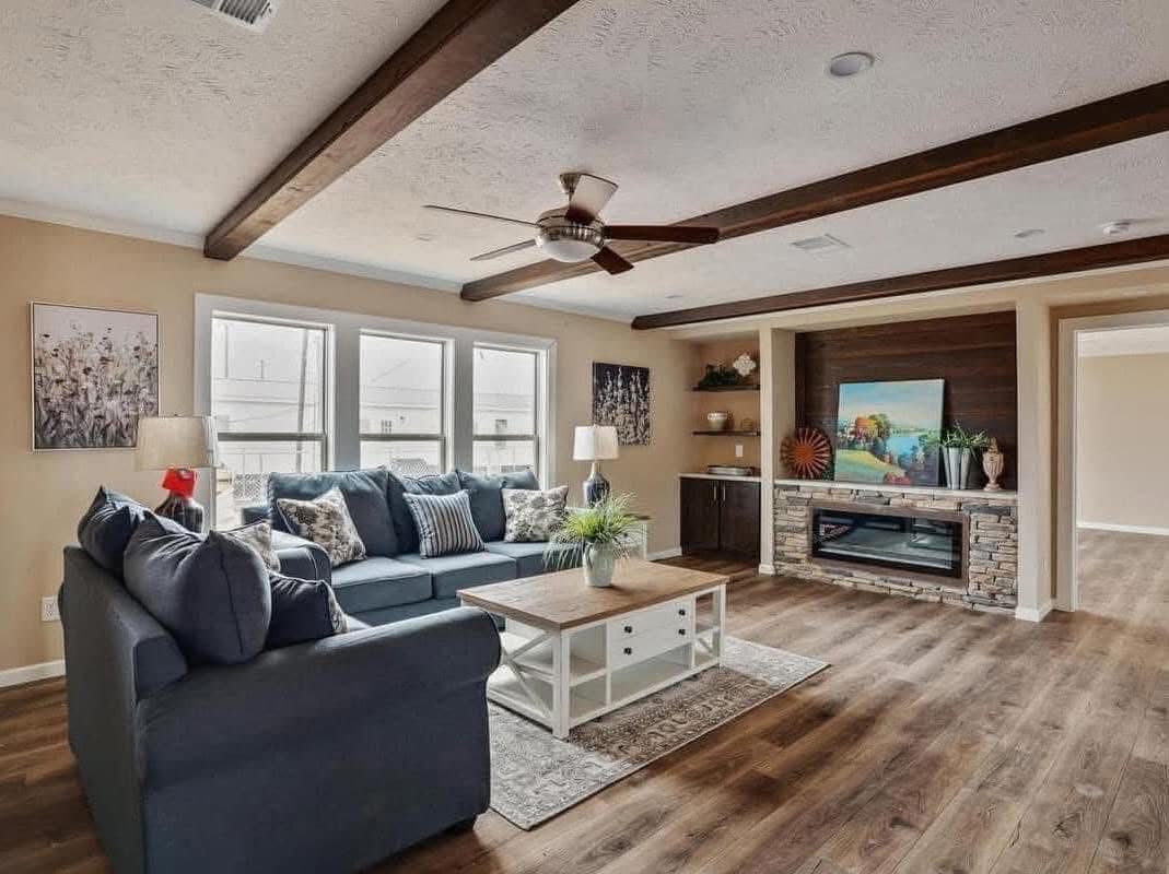 Cozy living room with a blue sectional sofa, striped cushions, and a wooden coffee table. Features a stone fireplace, ceiling fan, and natural light.