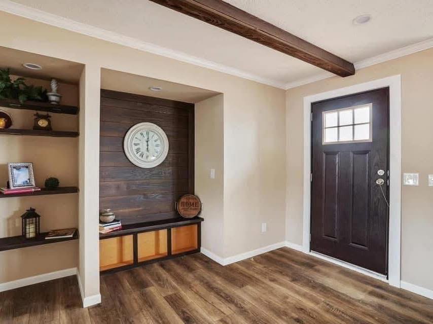 Cozy entryway with wood accents. Dark wood door, shelves with decor, clock on paneled wall, and warm flooring create an inviting, rustic feel.