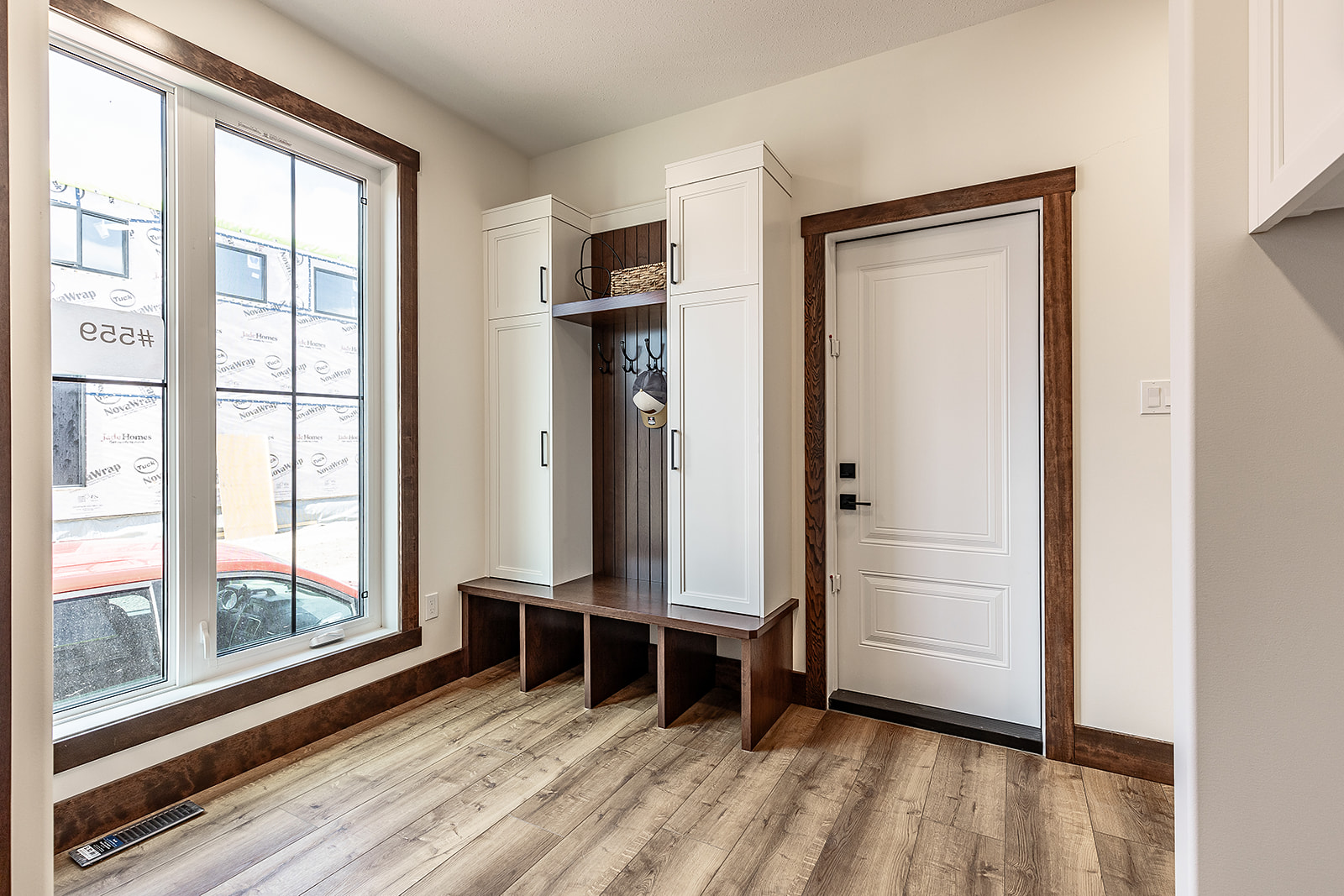 A bright entryway with light wood flooring, featuring a white door and a built-in storage bench with cabinets. Large window on the left. Cozy and organized.