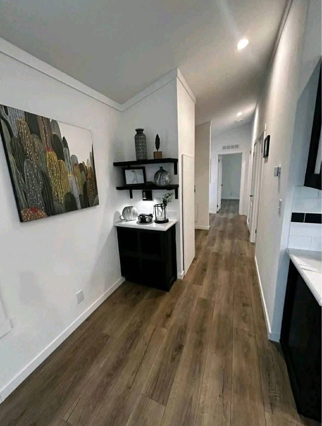 Modern hallway with wooden floors, white walls, and recessed lighting. Features cacti artwork, decorative shelves, and a cabinet, creating a cozy atmosphere.
