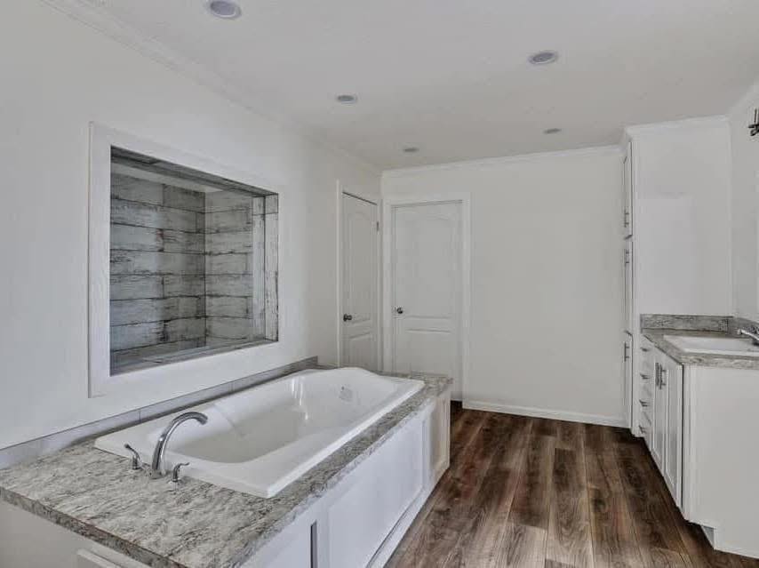 Modern bathroom with a large white tub featuring gray marble accents. The room has wood flooring, a wide mirror, and white cabinets, creating a serene atmosphere.
