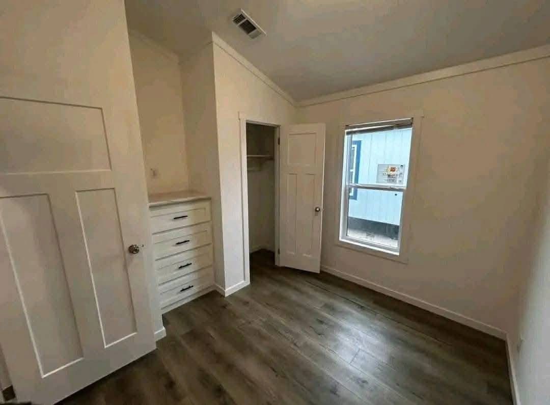 A small, empty room with light-colored walls and wood flooring. It features a window with partial blinds, a closet with a white door, and built-in drawers.