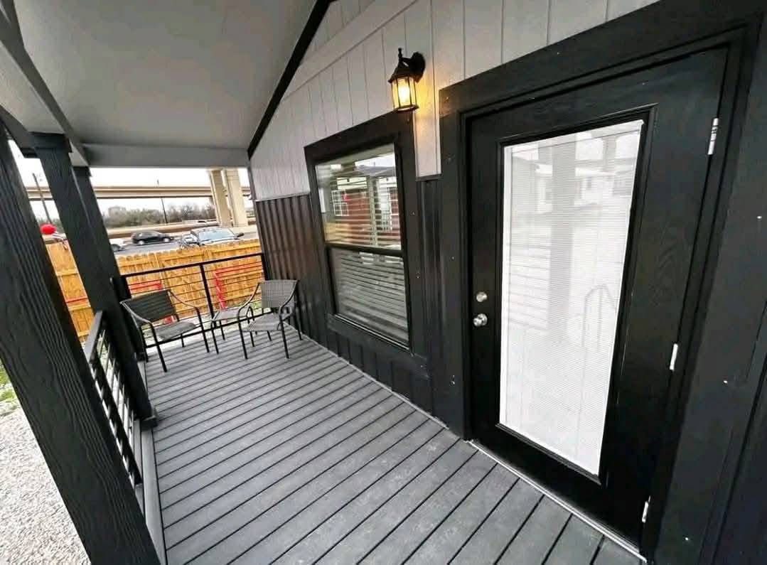 Covered porch with gray decking and black trim. Features a glass door, two chairs, a small table, and a wall-mounted lantern, creating a cozy and inviting atmosphere.